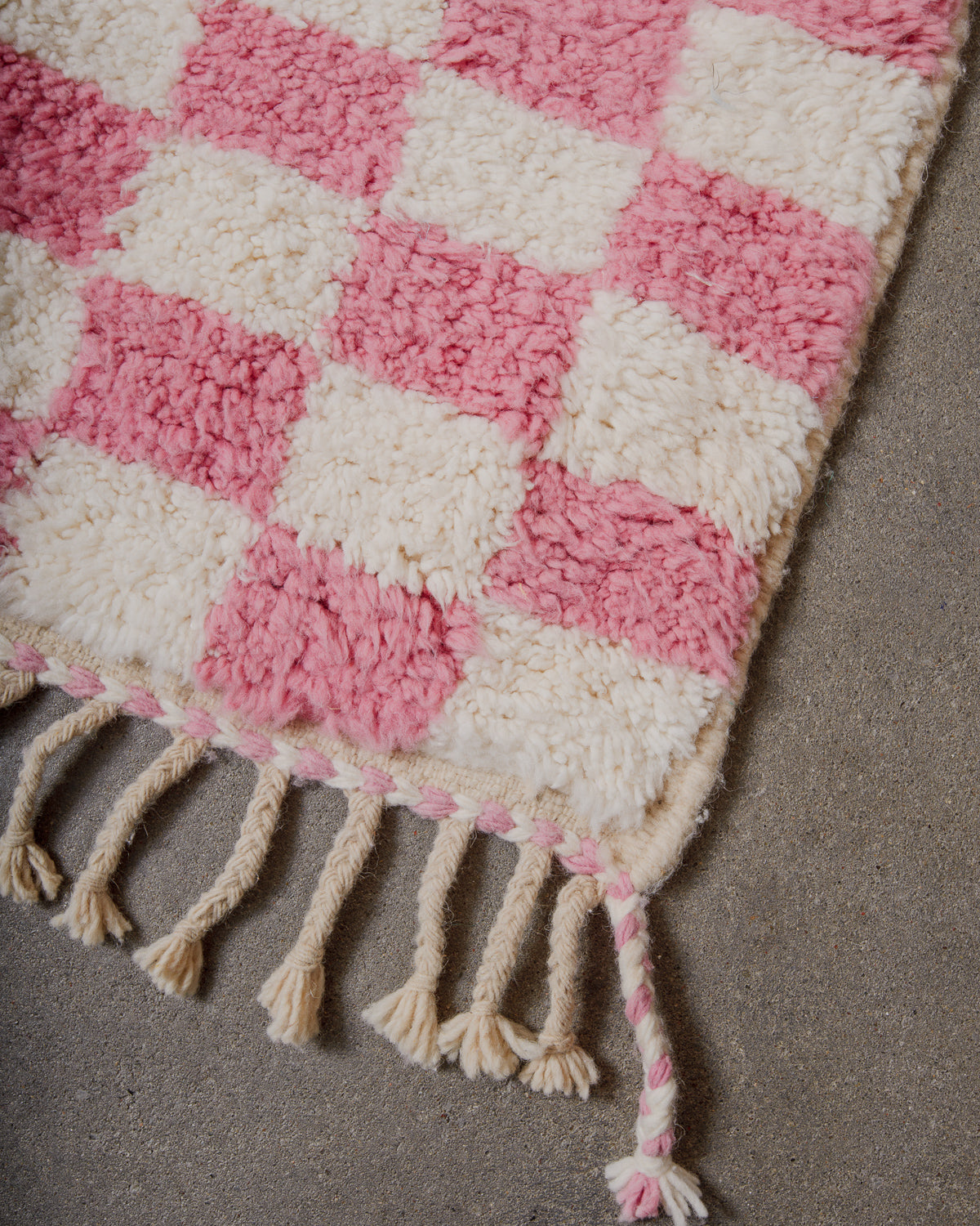 Corner of pink and cream checkered rug with natural wool tassels on concrete floor.